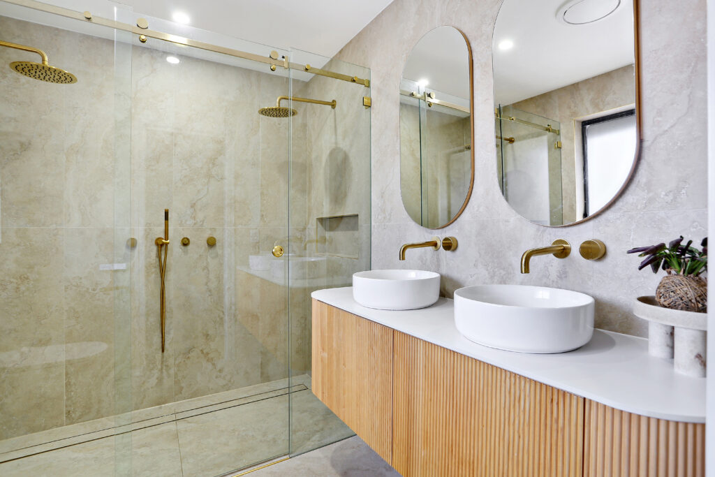 Double vanity with two vessel basins, fluted timber cabinet and arched mirrors beside a glass-screened shower